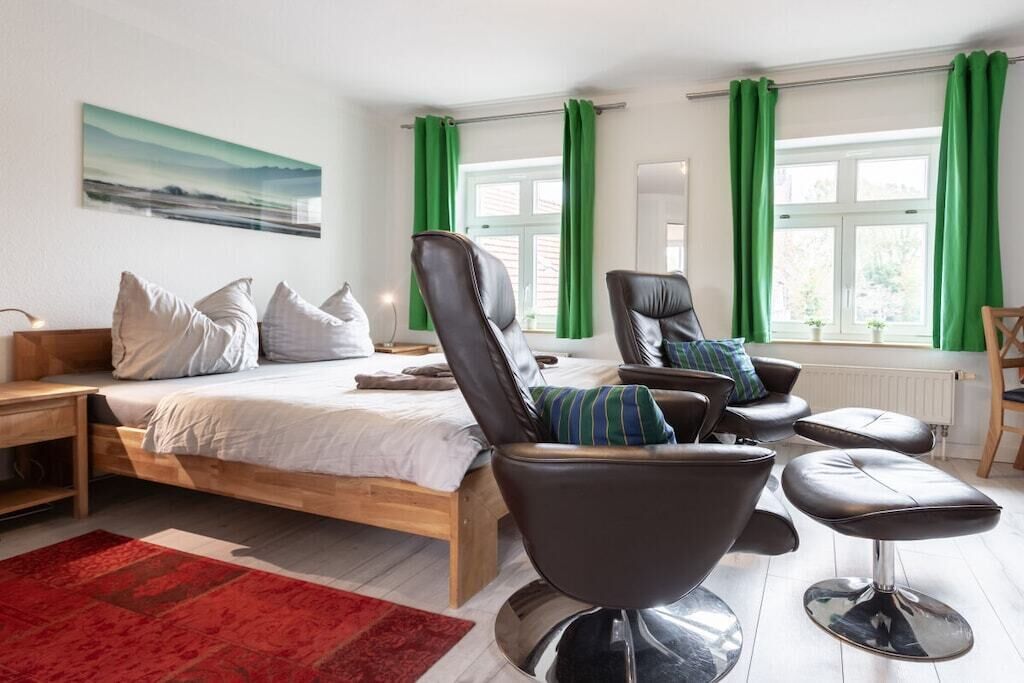 A bedroom with a bed, two chairs, a red rug, and a window with green curtains.