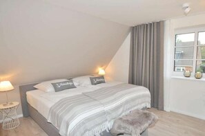 Un dormitorio con cama, lámparas de noche, una ventana con cortinas y una pequeña mesa con lámpara.
