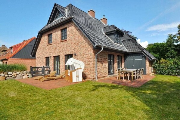 Una casa de dos pisos con fachada de ladrillo, techo inclinado y chimenea visible. Cuenta con un patio con mobiliario exterior y una zona cubierta.