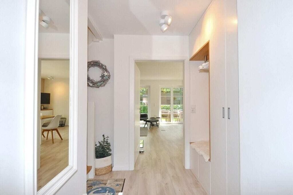 A hallway with a wooden floor, a white wall with a wreath decoration, and a door leading to a room with a bed and a window.
