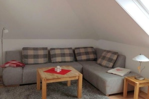 Ein gemütliches Schlafzimmer im Loftsstil mit schrägem Dach, einem grauen Sofa mit Karokissen, einem Holztisch und einer Nachttischlampe.