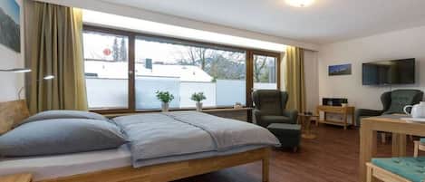 A room with a bed, a chair, a small table, a television, and a window with curtains.