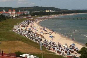 A beach with rows of beach chairs and umbrellas, a pier extending into the water, and a building with red roofs in the background. - Villa Bernstein Apt. 32 (Binz)