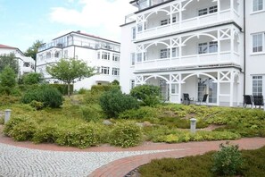Een meerlaags gebouw met balkons, omgeven door een aangelegde tuin met diverse struiken en een geplaveid pad.