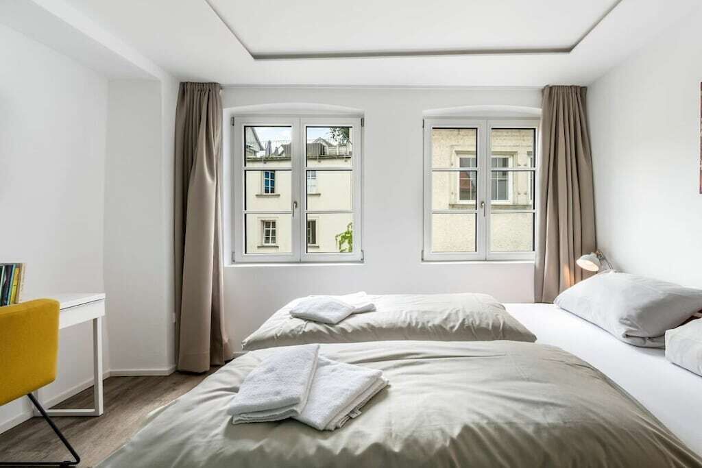 Ein Hotelzimmer mit zwei Betten, einem Schreibtisch und Blick auf Gebäude durch die Fenster.