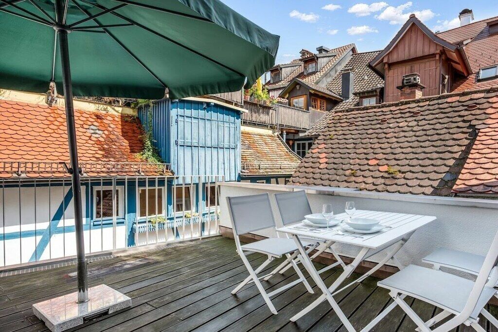 Een terras buiten met een tafel en stoelen, een groene parasol en een blauw gebouw op de achtergrond.