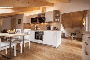 Une cuisine moderne avec des meubles blancs, un four encastré et un four à micro-ondes. On y trouve une table à manger avec des chaises et un panier de fruits.