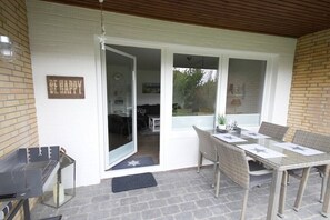 A covered patio with a table and chairs, a grill, and a door leading to an indoor area. - Holiday apartment with 2 bedrooms (Büsumer Deichhausen)