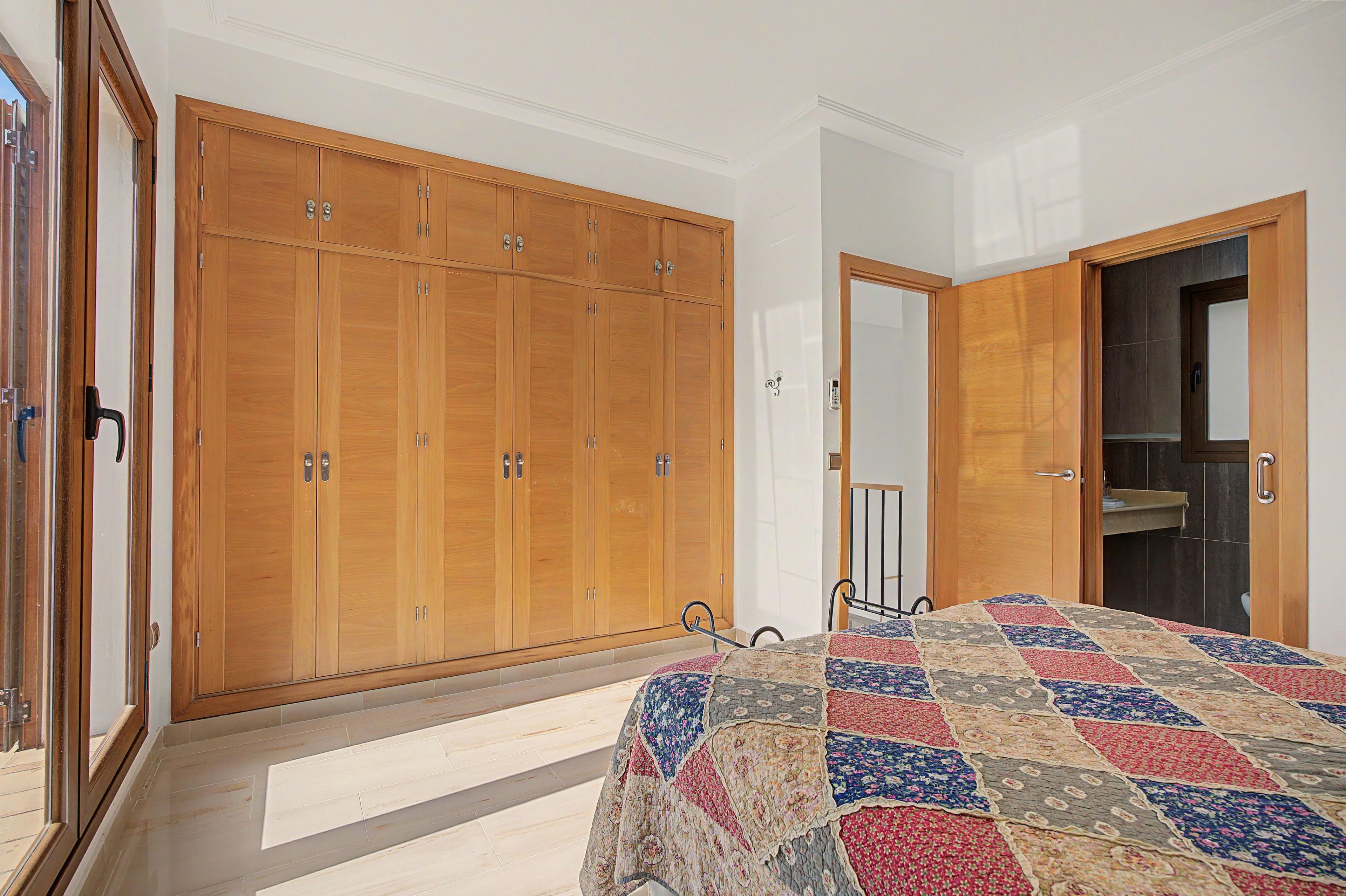 Un dormitorio con un armario grande, una cama con colcha estampada, y un baño visible a través de una puerta abierta.