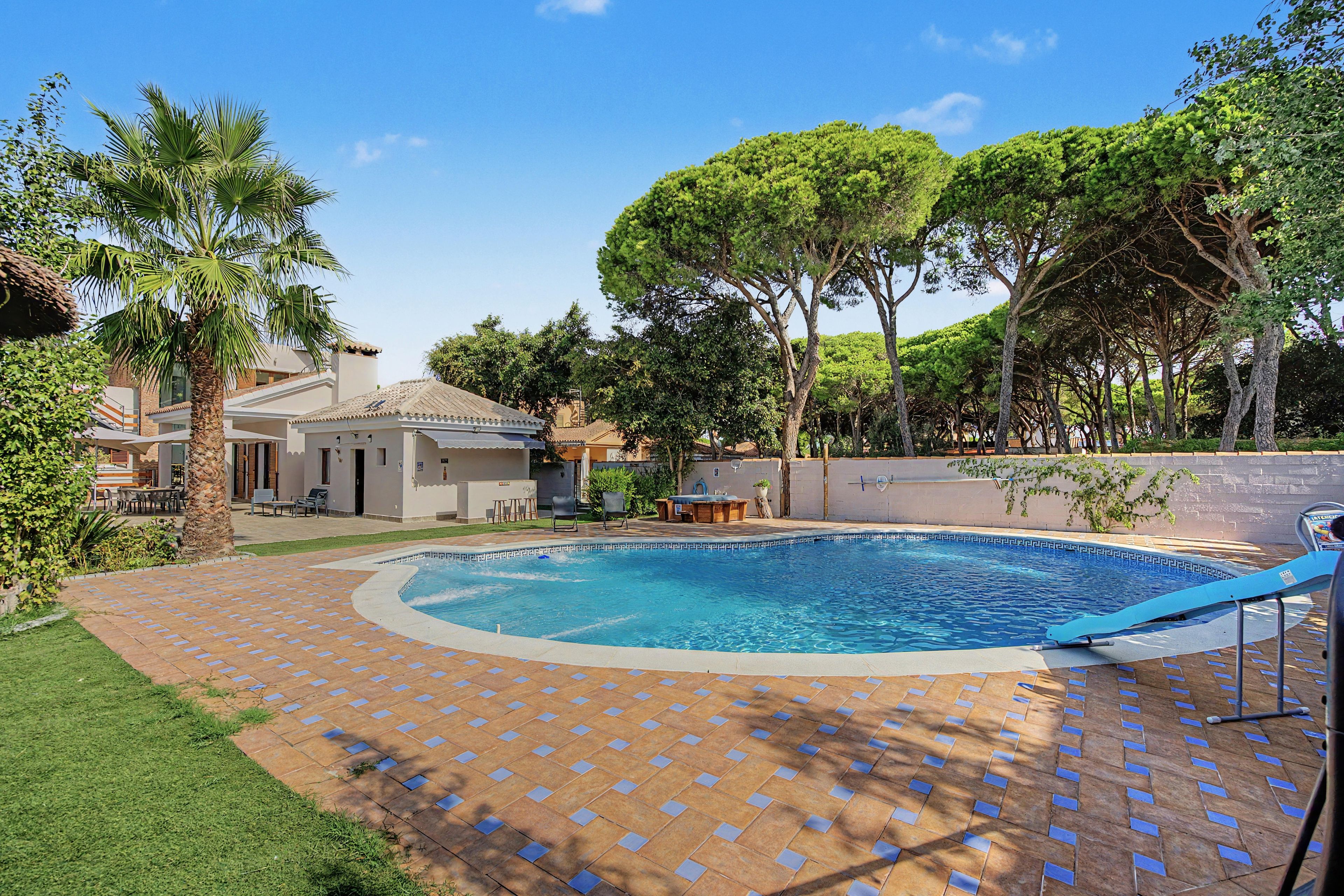 Una piscina con solárium de baldosas, rodeada de vegetación y un edificio con tejado de tejas.