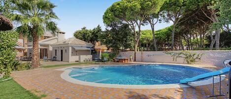 Una piscina con solárium de baldosas, rodeada de vegetación y un edificio con tejado de tejas.
