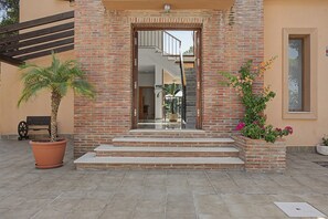 Edifici de maó amb entrada de vidre, escales d'accés, una banca i plantes en testos.