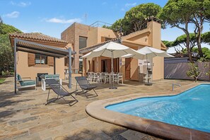 Un área de piscina con sillones de descanso, sombrillas y un edificio con tejado de tejas.