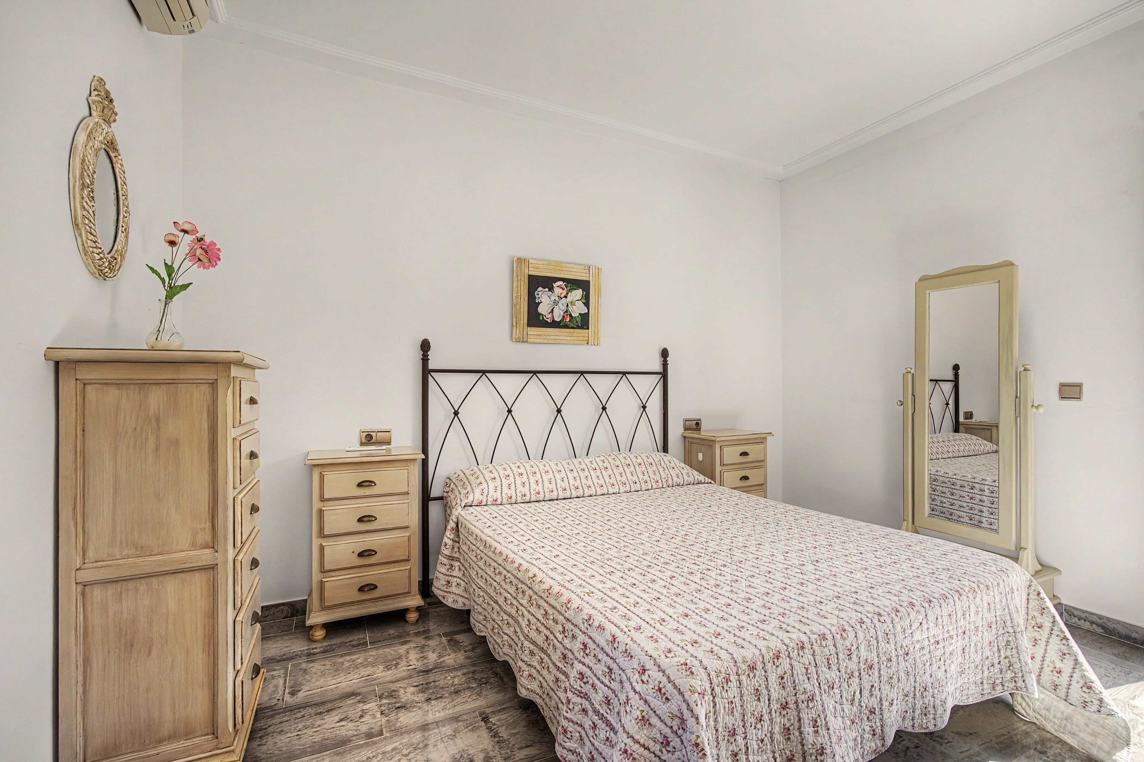 Une chambre à coucher comprenant un lit, une commode, des tables de chevet, un miroir et un tableau floral accroché au mur.