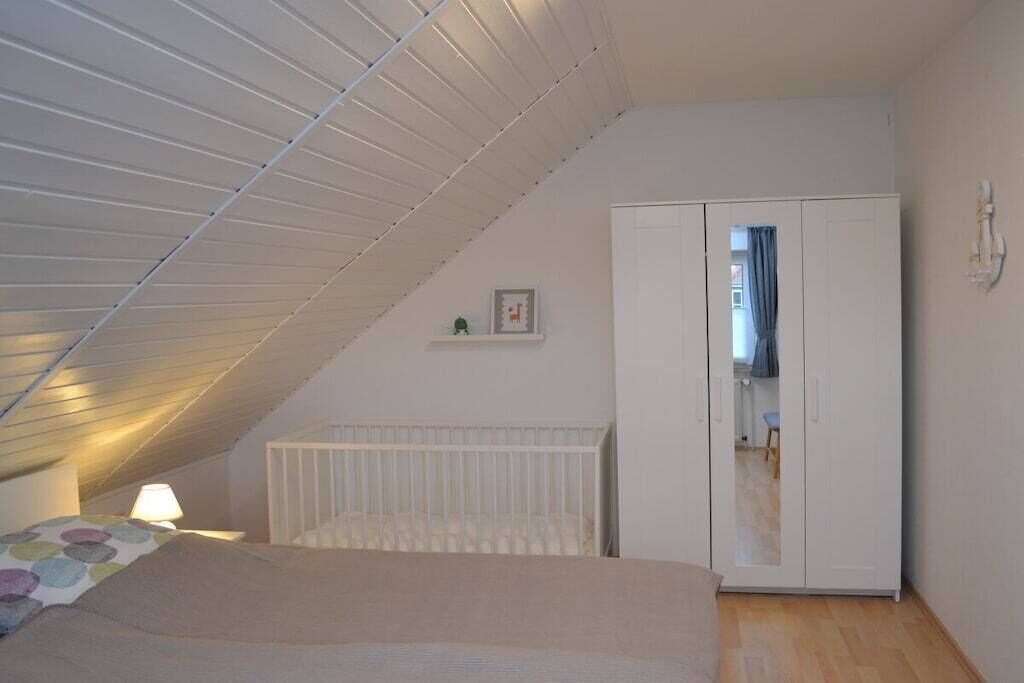 A bedroom with a sloped ceiling, a bed, a radiator, a wardrobe, and a small shelf with a picture.