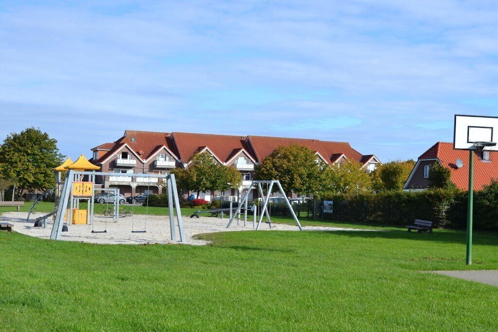 A playground with swings and a climbing structure, surrounded by a grassy area and residential buildings in the background.