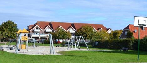 A playground with swings and a climbing structure, surrounded by a grassy area and residential buildings in the background.