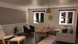 A living room with a sofa, armchair, coffee table, and a small table with a lamp. There are windows with curtains, a hanging light fixture, and a plant on the windowsill. - B and B in Bjuråker with swimming pool (Bjuråker)