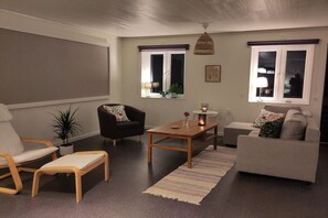 A living room with a sofa, armchair, coffee table, and a small table with a lamp. There are windows with curtains, a hanging light fixture, and a plant on the windowsill.