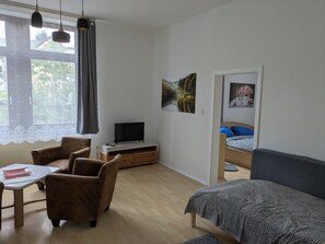 A room with a wooden floor, a desk with a chair, a bed, a television on a wooden stand, and a window with curtains.