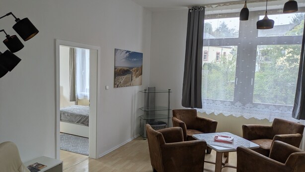 Ein Zimmer mit einem Bett, einem Schreibtisch, einem Sessel, einem Tisch und einem Vorhangfenster.