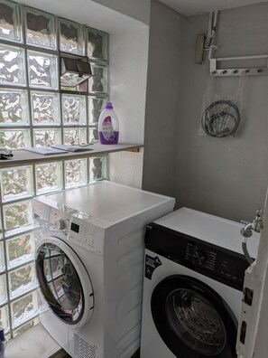 A laundry room with a front-loading washer and dryer, a shelf with a bottle of Toptil detergent, and a window with a view of trees.