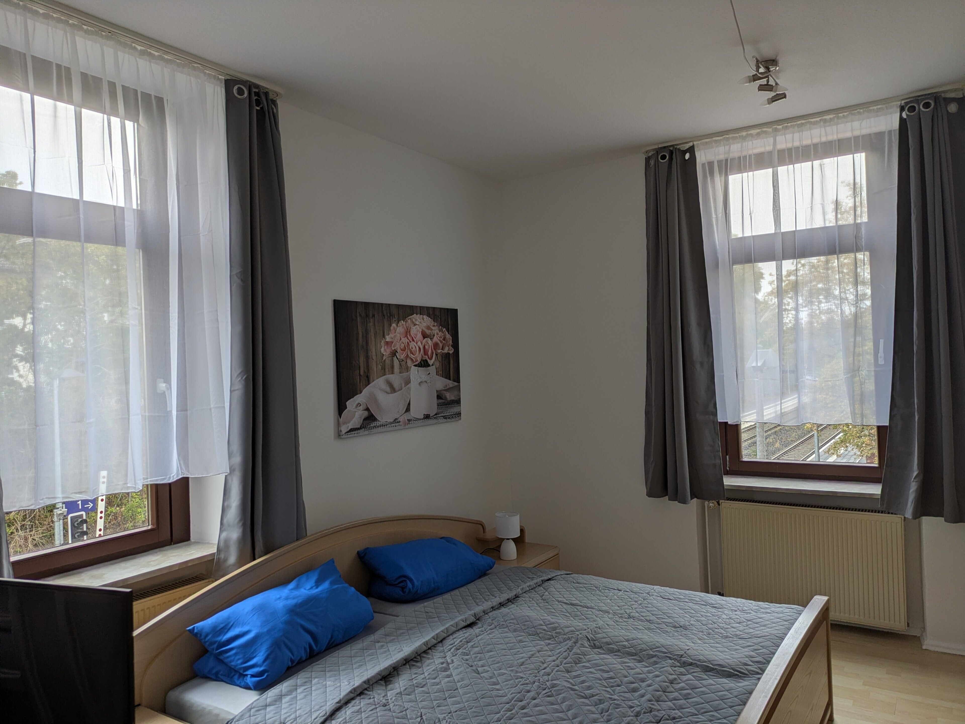 Ein Schlafzimmer mit Bett, Kissen, einem Nachttisch, einer Lampe, einem Gemälde an der Wand und Fenstern mit Vorhängen.