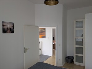 A room with a bed, a door leading to another room, a wall-mounted picture, and a light fixture.