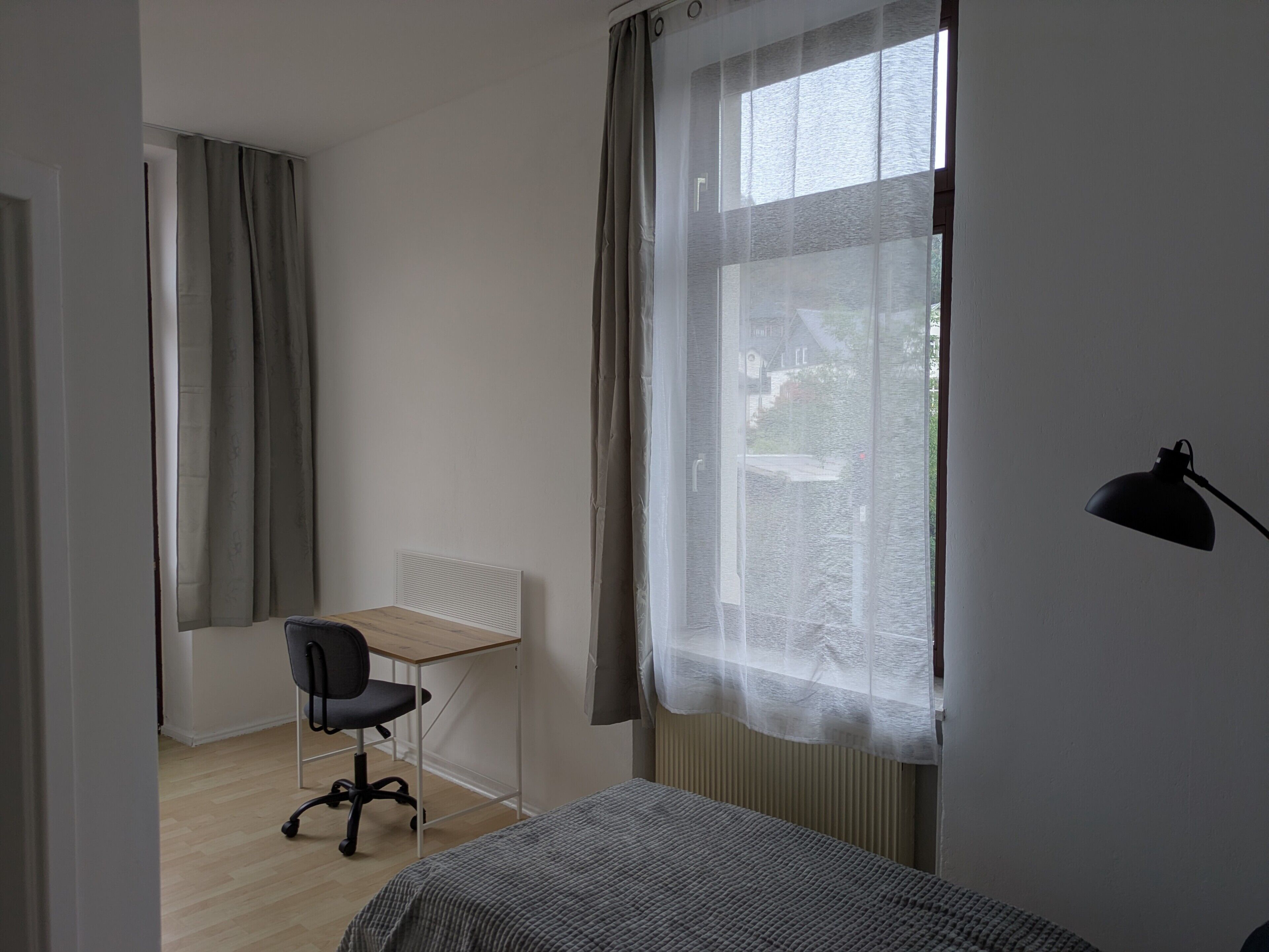 Ein Zimmer mit einem Bett, einem Schreibtisch, einem Stuhl und einem fensterbepflanzten Fenster.