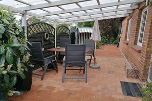 Een overdekt terras met een tafel, stoelen en een strepenpatroon parasol, met een bakstenen muur.