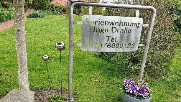 Et metalskilt med tekst, der angiver en ejendom ved navn 'Ferienwohnung Ingo Dralle' og et telefonnummer.