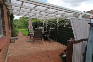 A covered patio area with brick flooring, two wicker chairs, and a table. - Anke Dralle Strandweg 11 (Elbinsel Krautsand)