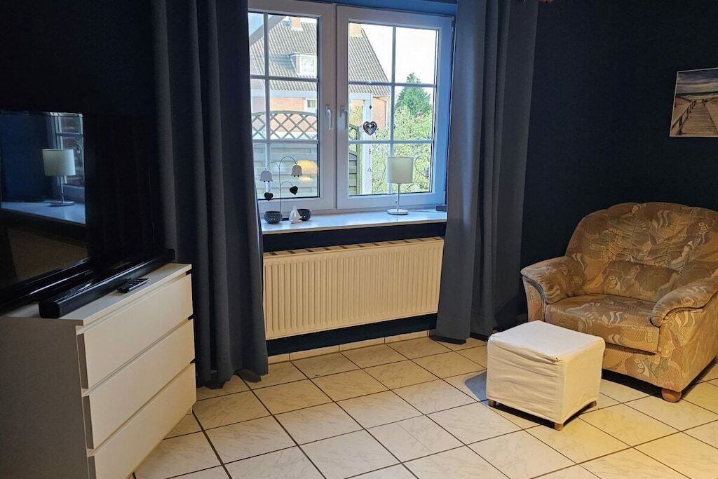Una habitación con ventana, radiador, televisor, una silla y una mesita.