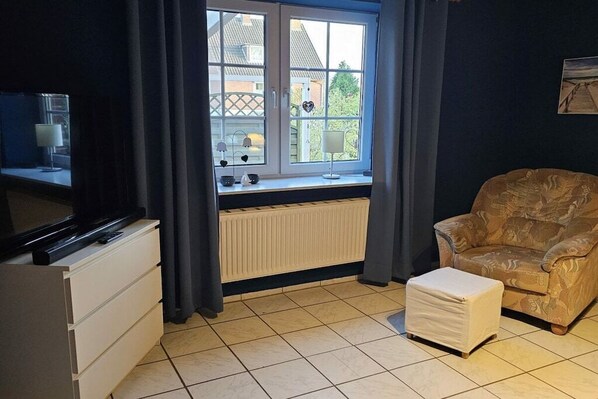 Ein Zimmer mit Fenster, Heizkörper, Fernseher, Stuhl und kleinem Tisch.