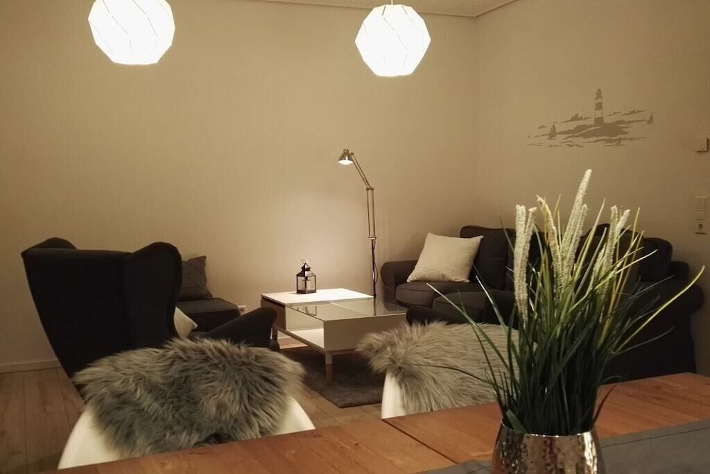 A modern living room with a sofa, armchair, and a coffee table. There is a lamp, a vase with plants, and a wall art piece.