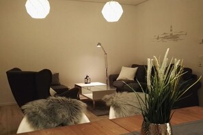 A modern living room with a sofa, armchair, and a coffee table. There is a lamp, a vase with plants, and a wall art piece. - Semi-detached house "LUV" (Butjadingen)