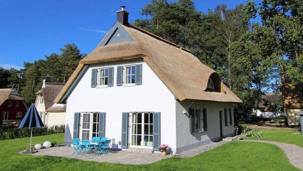 Uma casa com telhado de palha, varanda frontal e um pequeno jardim com móveis de exterior.