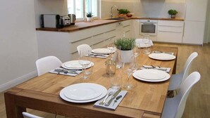 Uma sala de jantar com mesa de madeira posta para quatro pessoas, cadeiras brancas e uma cozinha ao fundo.