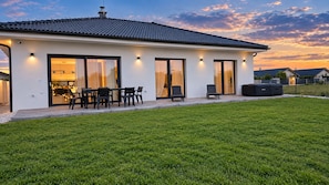 Una casa moderna con tetto piatto, ampie porte scorrevoli in vetro, un patio con mobili da esterno e un prato curato.