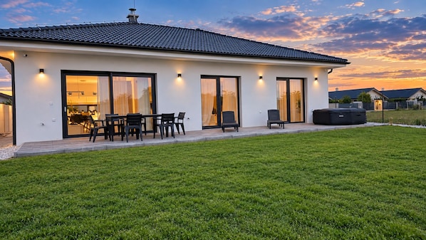 A modern house with a flat roof, large sliding glass doors, a patio with outdoor furniture, and a well-maintained lawn.