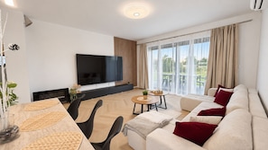 A modern living room with a flat-screen TV, a sofa set, a dining table with chairs, and a balcony with curtains.