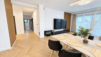 A modern living room with a large flat-screen TV, a dining area with a wooden table and black chairs, and a view of the outdoors through large windows.