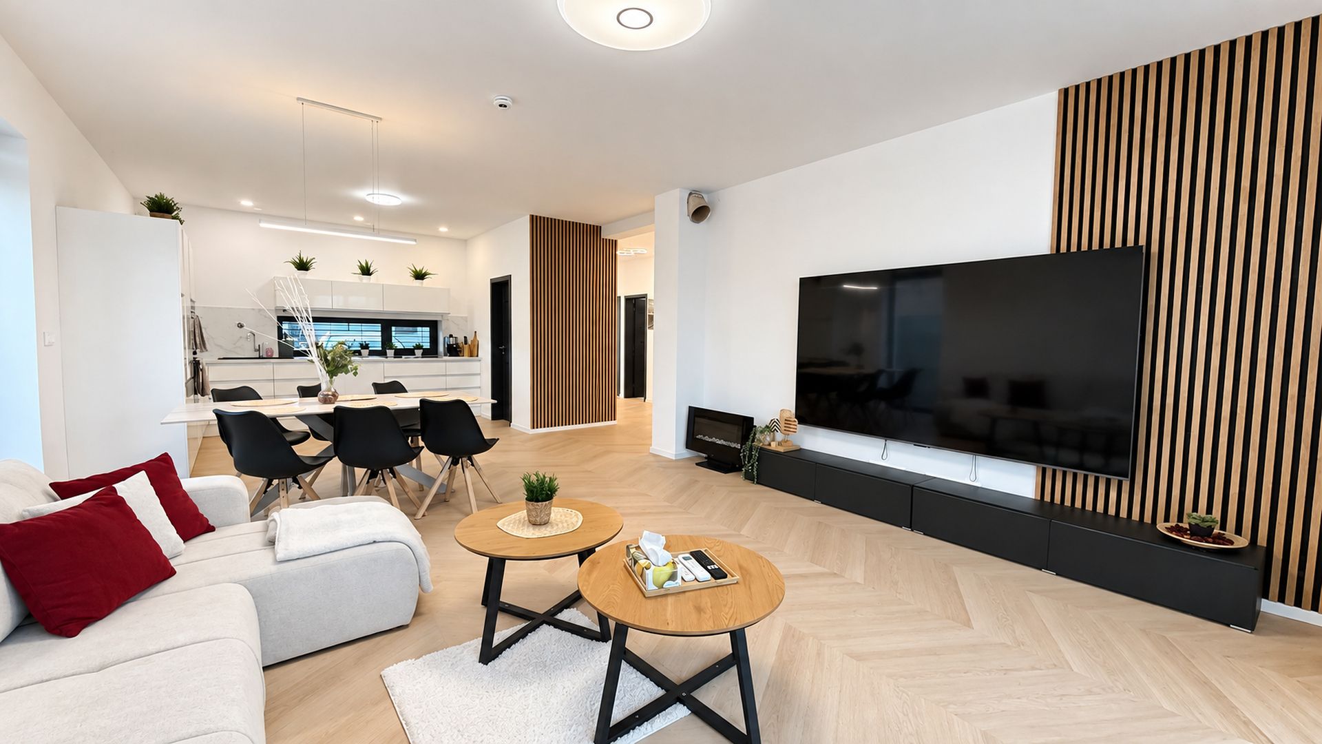 A modern living room with a flat-screen TV, a sofa, two coffee tables, and a kitchen area in the background.