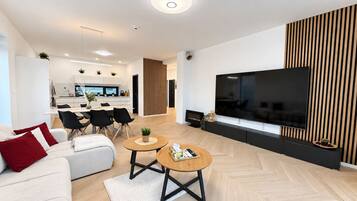 A modern living room with a flat-screen TV, a sofa, two coffee tables, and a kitchen area in the background.