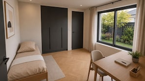 A bedroom with a bed, a desk, and a window with a view of greenery.