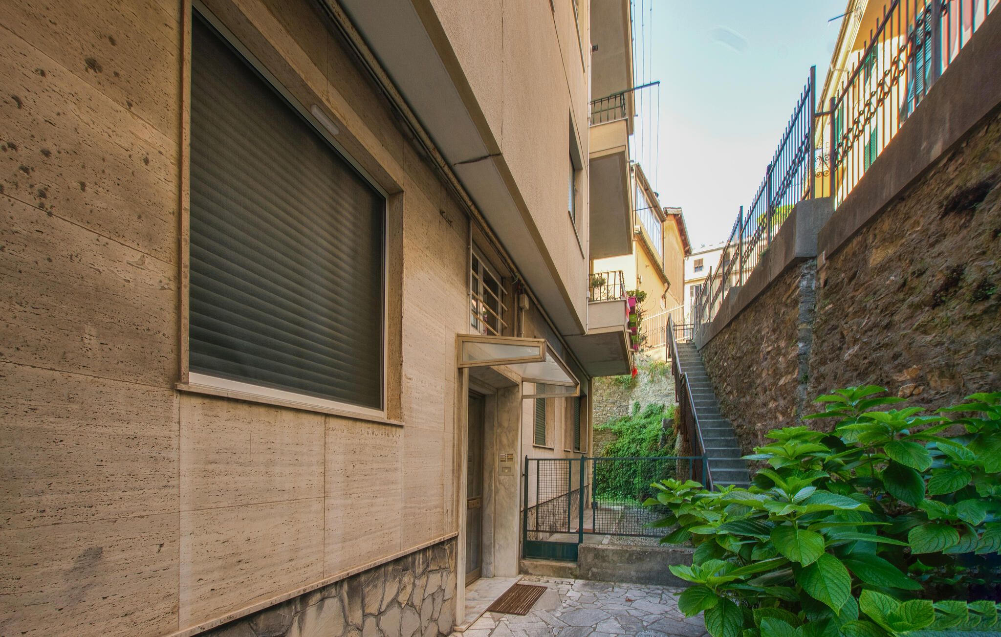 Un callejón estrecho con una pared de piedra a un lado y un edificio con un toldo cerrado al otro. Una escalera conduce a un balcón con barandilla de metal.