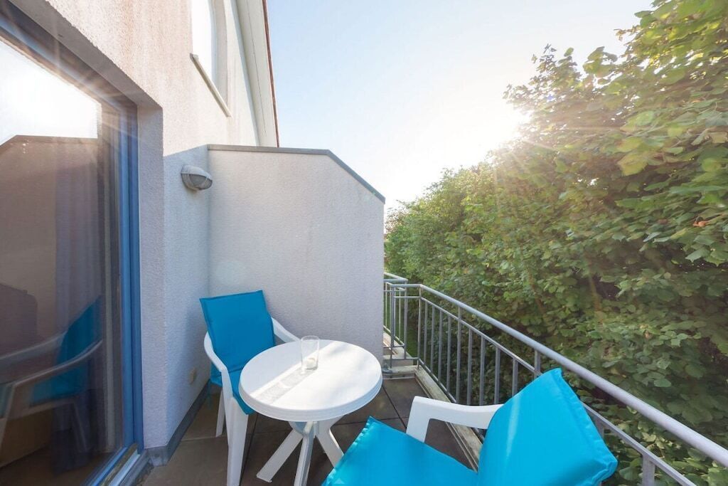 A balcony with a white table and blue chairs, a glass door, and a view of greenery.