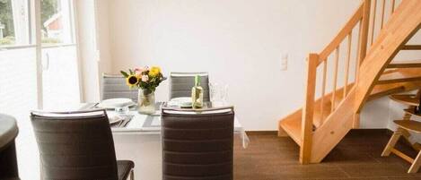 A dining area with a table set for two, two chairs, a vase with flowers, and a wooden staircase.
