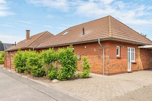 Une maison à deux étages, construite en briques, avec un toit en tuiles, une cheminée et un petit jardin agrémenté d’arbustes.