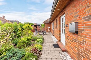 Une maison en briques avec un jardin, une terrasse avec une table et des chaises, et une boîte aux lettres fixée au mur.
